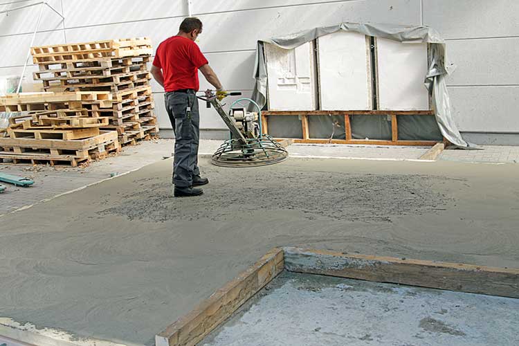 Foto: Eine Person in Arbeitskleidung bedient eine Glättmaschine auf einer frisch betonierten Fläche. 