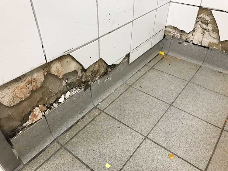 Foto: Gefliester Wand- und Bodenbereich in einer Ecke, in dem mehrere Wandfliesen beschädigt oder komplett entfernt sind. Darunter sind bröckelnder Putz und freigelegte, unregelmäßige Mörtelschichten erkennbar.  