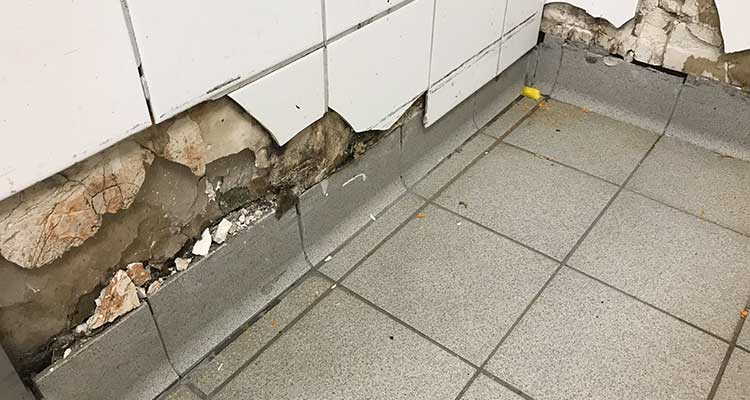 Foto: Gefliester Wand- und Bodenbereich in einer Ecke, in dem mehrere Wandfliesen beschädigt oder komplett entfernt sind. Darunter sind bröckelnder Putz und freigelegte, unregelmäßige Mörtelschichten erkennbar.  