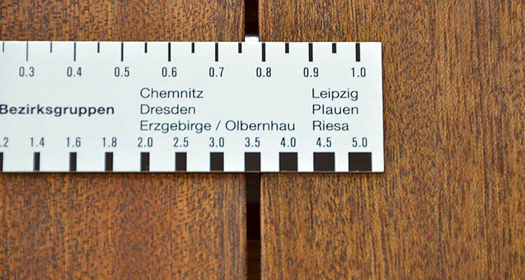 Foto: Gefliester Wand- und Bodenbereich in einer Ecke, in dem mehrere Wandfliesen beschädigt oder komplett entfernt sind. Darunter sind bröckelnder Putz und freigelegte, unregelmäßige Mörtelschichten erkennbar.  