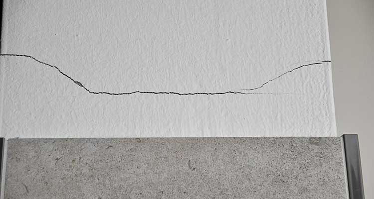 Foto: Gefliester Wand- und Bodenbereich in einer Ecke, in dem mehrere Wandfliesen beschädigt oder komplett entfernt sind. Darunter sind bröckelnder Putz und freigelegte, unregelmäßige Mörtelschichten erkennbar.  