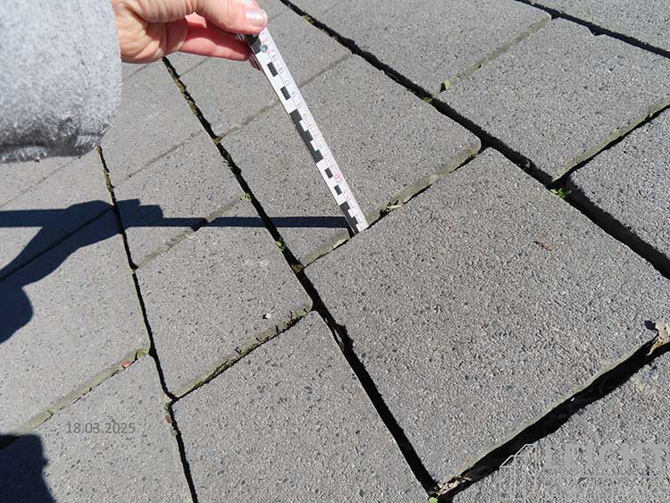 Das Bild zeigt einen Pflasterbelag, bei dem mittels eines Meterstabs die Tiefe der Fugenverfüllung gemessen wird. 