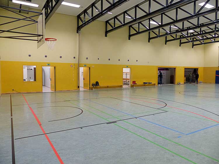 Sporthalle mit gelben Wänden und einer Spielfläche aus bläulich grauem Bodenbelag, markiert mit Linien für unterschiedliche Sportarten in Schwarz, Rot, Grün und Blau. Oben links hängt ein Basketballkorb. Entlang der Wand sind mehrere offene Türen und Notausgänge erkennbar. Decke mit schwarzen Stahlträgern und Beleuchtung. 