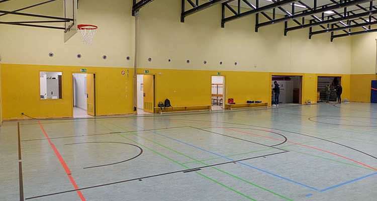 Sporthalle mit gelben Wänden und einer Spielfläche aus bläulich grauem Bodenbelag, markiert mit Linien für unterschiedliche Sportarten in Schwarz, Rot, Grün und Blau. Oben links hängt ein Basketballkorb. Entlang der Wand sind mehrere offene Türen und Notausgänge erkennbar. Decke mit schwarzen Stahlträgern und Beleuchtung.