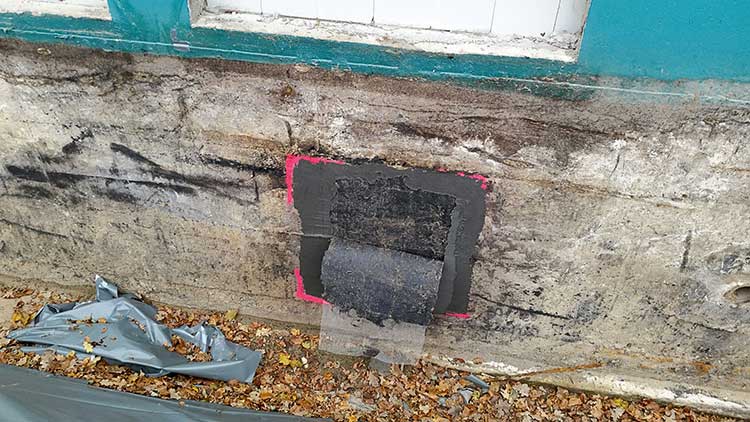 Mauer einer Gebäudefassade mit beschädigtem Putz nahe einem Fensterrahmen. Mittig ist ein rechteckiger, dunkler Bereich sichtbar, der provisorisch mit Dichtmasse beschichtet und mit einer schwarzen Isolierbahn abgedeckt ist. Ein pinker Rahmen markiert den Arbeitsbereich. Im Vordergrund liegen Herbstblätter und Reste einer grauen Abdeckplane.