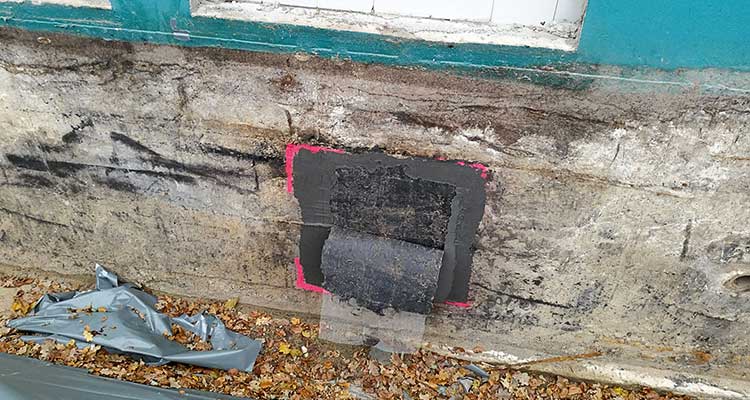 Mauer einer Gebäudefassade mit beschädigtem Putz nahe einem Fensterrahmen. Mittig ist ein rechteckiger, dunkler Bereich sichtbar, der provisorisch mit Dichtmasse beschichtet und mit einer schwarzen Isolierbahn abgedeckt ist. Ein pinker Rahmen markiert den Arbeitsbereich. Im Vordergrund liegen Herbstblätter und Reste einer grauen Abdeckplane. 