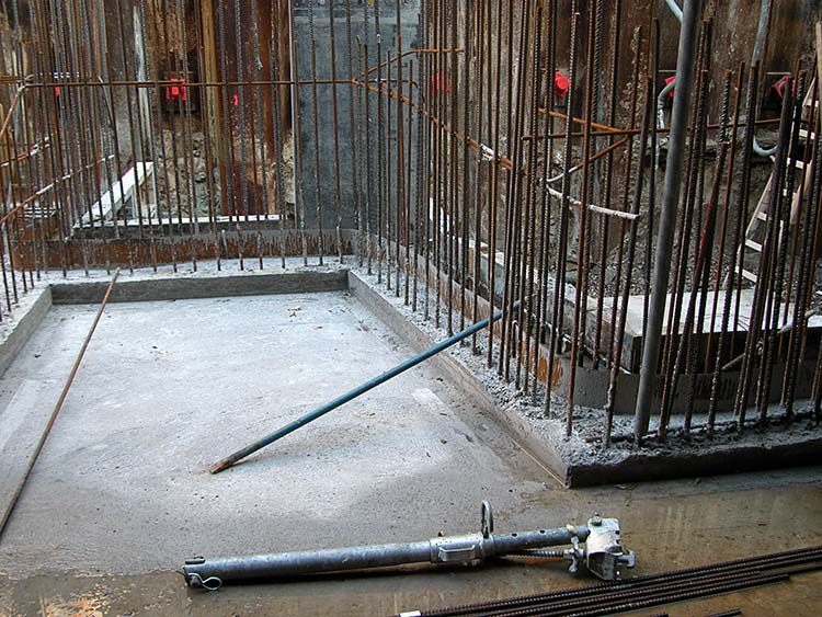 Baustelle mit frischem Betonfundament, dicht stehenden senkrechten Bewehrungsstäben und einem auf dem Boden liegenden Metallrohr 
