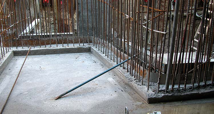 Baustelle mit frischem Betonfundament, dicht stehenden senkrechten Bewehrungsstäben und einem auf dem Boden liegenden Metallrohr 