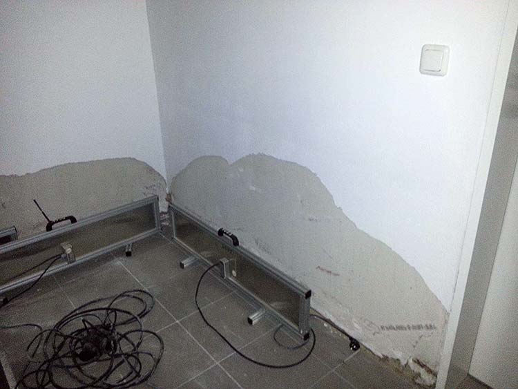 Innenwand mit feuchter, abgeplatzter Putzfläche, davor Metallgestelle und Kabel auf einem gefliesten Boden. 