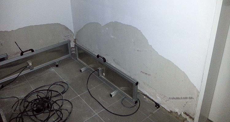 Innenwand mit feuchter, abgeplatzter Putzfläche, davor Metallgestelle und Kabel auf einem gefliesten Boden. 