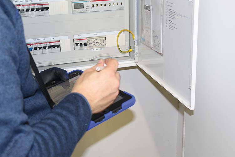 Person prüft mit Tablet einen geöffneten Elektroschaltkasten mit Sicherungen und Messgeräten. 