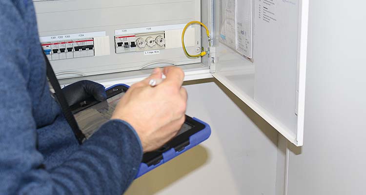 Person prüft mit Tablet einen geöffneten Elektroschaltkasten mit Sicherungen und Messgeräten. 