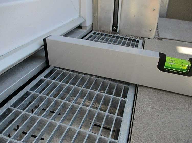 Eine Wasserwaage liegt auf einem Gitterrost, der sich zwischen einer Türschwelle und einem betonierten Bereich befindet. Es ist deutlich zu sehen, dass die Türschwelle höher liegt als der Gitterrost und der anschließende Bereich. 