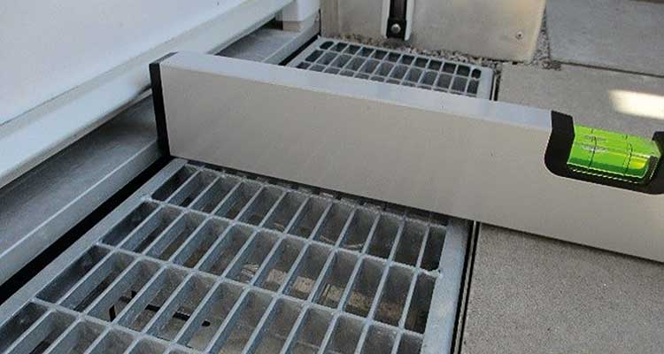 Eine Wasserwaage liegt auf einem Gitterrost, der sich zwischen einer Türschwelle und einem betonierten Bereich befindet. Es ist deutlich zu sehen, dass die Türschwelle höher liegt als der Gitterrost und der anschließende Bereich. 