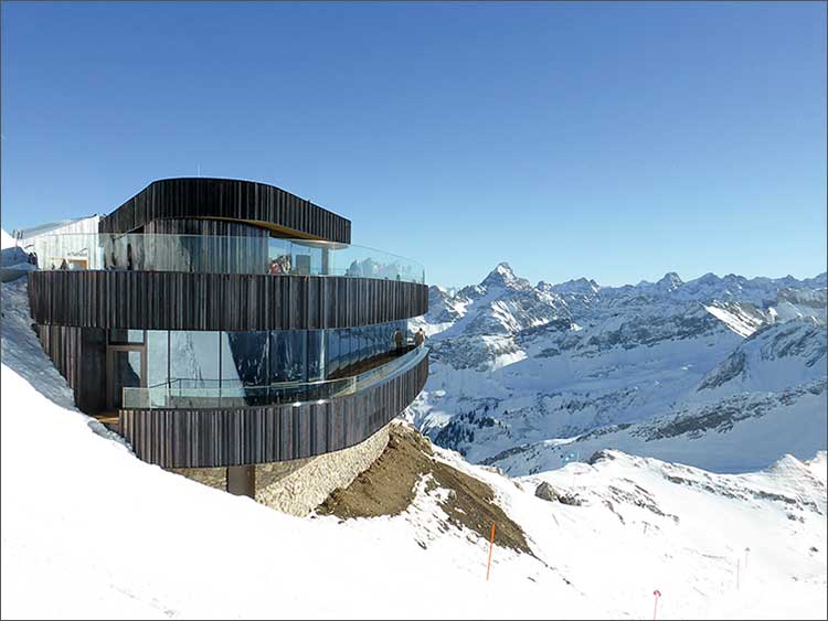 Modernes Gebäude aus dunklem Holz und Glas mit zwei geschwungenen Etagen, das an einem verschneiten Berghang liegt. 