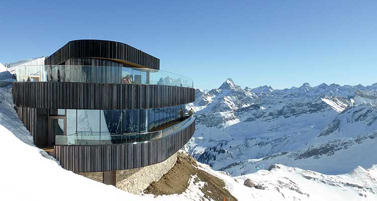 Modernes Gebäude aus dunklem Holz und Glas mit zwei geschwungenen Etagen, das an einem verschneiten Berghang liegt. 