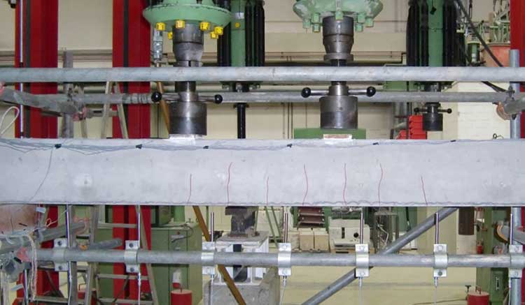 Betonbalken in einer strukturellen Belastungsprüfung. Eine hydraulische Presse drückt auf den Balken. Der Balken ist horizontal eingespannt, mit sichtbaren Rissen entlang seiner Unterseite. 