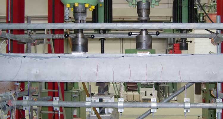 Betonbalken in einer strukturellen Belastungsprüfung. Eine hydraulische Presse drückt auf den Balken. Der Balken ist horizontal eingespannt, mit sichtbaren Rissen entlang seiner Unterseite. 