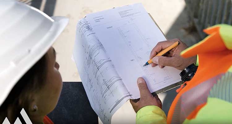 Eine Person hält ein Klemmbrett mit technischen Bauplänen. 