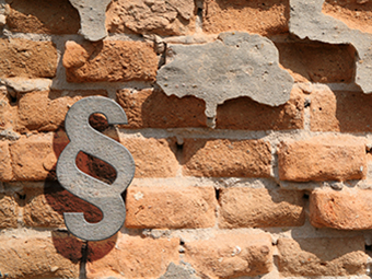 Backsteinmauer mit partiell abbröckelndem Putz; links unten hängt ein Paragraphensymbol aus Metall, das Schatten auf die Mauer wirft. Der sichtbare Putz ist ungleichmäßig verteilt und zeigt graue Farbflächen, die mit den Backsteinen kontrastieren. 