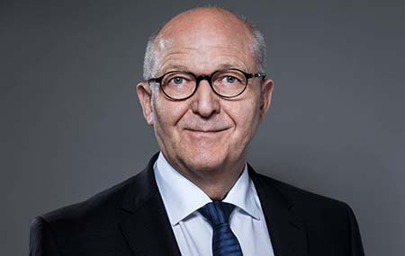 Porträt eines älteren Mannes mit heller, teilweise kahler Kopfhaut. Er trägt eine runde schwarze Brille, einen dunklen Anzug, ein weißes Hemd und eine dunkelblaue Krawatte. Frontalaufnahme vor einfarbig grauem Hintergrund. 