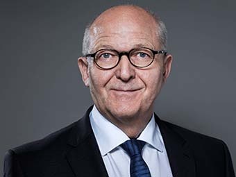 Porträt eines älteren Mannes mit heller, teilweise kahler Kopfhaut. Er trägt eine runde schwarze Brille, einen dunklen Anzug, ein weißes Hemd und eine dunkelblaue Krawatte. Frontalaufnahme vor einfarbig grauem Hintergrund. 