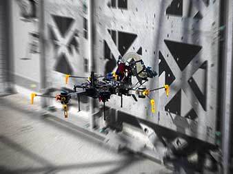 Das Bild zeigt eine Drohne an der Testwand im DroneHub mit modularen Bauelementen für Versuche mit fliegenden Baurobotern. 