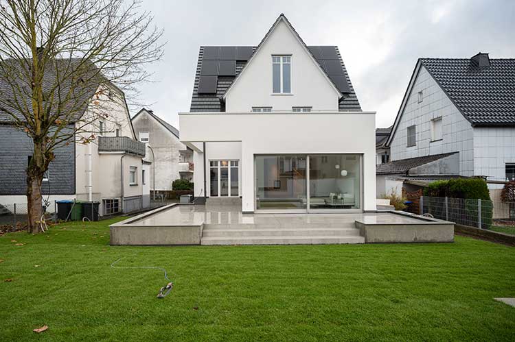 Das Bild zeigt ein modernisiertes Haus mit spitzem Giebel, PV-Anlage auf dem Dach, einem Anbau mit großen Glasschiebetüren und einer Terrasse mit hoch glänzendem Steinbelag, vom Garten aus gesehen. 