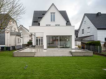 Das Bild zeigt ein modernisiertes Haus mit spitzem Giebel, PV-Anlage auf dem Dach, einem Anbau mit großen Glasschiebetüren und einer Terrasse mit hoch glänzendem Steinbelag, vom Garten aus gesehen. 