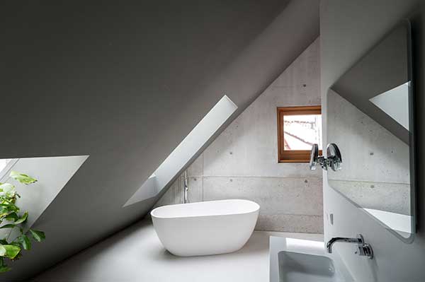 Das Bild zeigt ein modernes Badezimmer mit Dachschräge vom Boden bis zur Decke, rohen Betonwänden, in der Höhe versetzten Fenstern und einer frei stehenden weißen Badewanne.