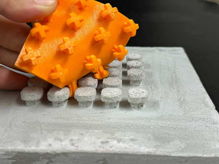 Das Bild zeigt ein Modell des Klettverschlusses zur Verbindung von Gebäudeteilen. Ein Teil ist eine graue Betonplatte mit pilzförmigen Noppen, der Gegenpart ist aus orangefarbigem Kunststoff und hat 