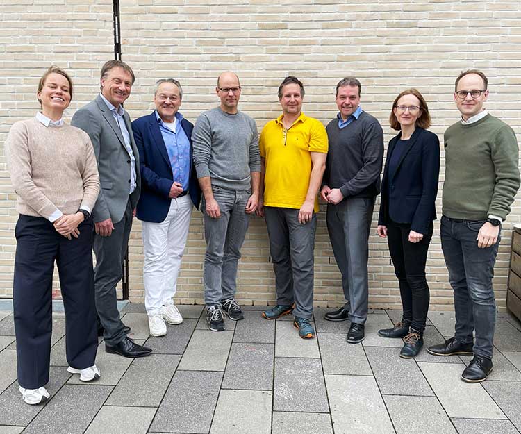 Gruppenfoto von acht Personen, die nebeneinander vor einer hellbeigen Ziegelwand stehen. Verschiedene Kleidungsstile: Anzüge, Freizeitkleidung und ein gelbes Polohemd. Unter ihnen befinden sich sowohl Frauen als auch Männer. Der Boden besteht aus großflächigen, grauen Pflastersteinen. 