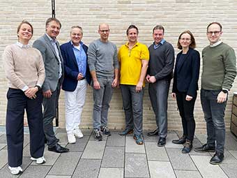 Gruppenfoto von acht Personen, die nebeneinander vor einer hellbeigen Ziegelwand stehen. Verschiedene Kleidungsstile: Anzüge, Freizeitkleidung und ein gelbes Polohemd. Unter ihnen befinden sich sowohl Frauen als auch Männer. Der Boden besteht aus großflächigen, grauen Pflastersteinen. 