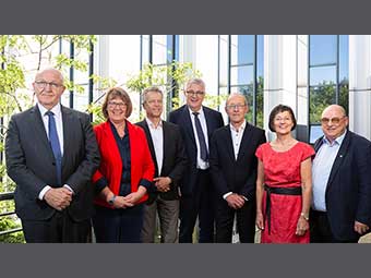 Eine Gruppe von sieben Personen – fünf Männer und zwei Frauen – in formeller Kleidung steht in zwei Reihen vor einem modernen Bürogebäude mit großen Glasfenstern. Im Hintergrund sind grüne Bäume zu sehen.  
