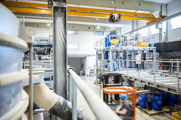 Industriehalle mit Hochregalen, in denen Fässer und andere Behälter stehen, sowie einer Hebeeinrichtung an einer Schiene, die an einem Stahlträger an der Decke befestigt ist. 