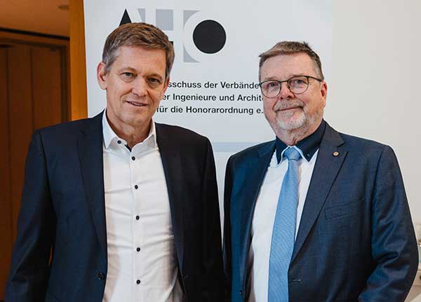 Zwei Männer in Anzügen stehen vor einem Roll-up mit dem Logo 