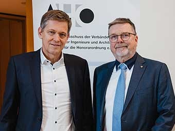 Zwei Männer in Anzügen stehen vor einem Roll-up mit dem Logo "AHO" und dem Text "Ausschuss der Verbände und Kammern der Ingenieure und Architekten für die Honorarordnung e.V.".  