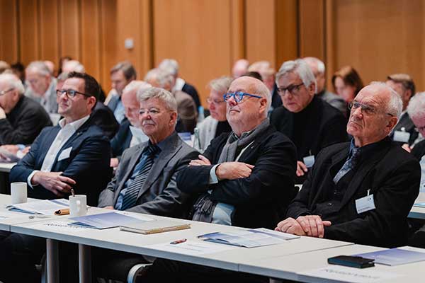 Mehrere Personen sitzen in einem Konferenzraum an Tischen in parallelen Reihen. Auf den Tischen liegen Broschüren, Notizbücher und Stifte. 
