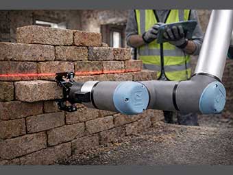 Ein Roboterarm hält einen Backstein, der halb in einer niedrigen Backsteinmauer steckt. 