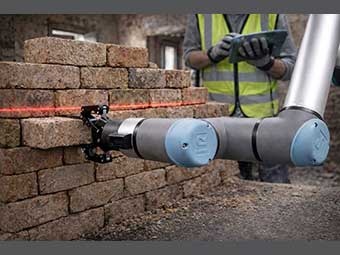 Ein Roboterarm hält einen Backstein, der halb in einer niedrigen Backsteinmauer steckt. 