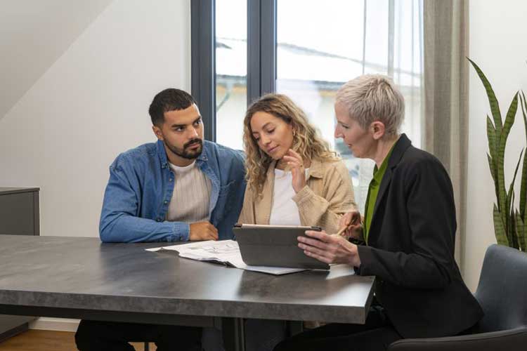 Foto: Drei Personen sitzen an einem Tisch, auf dem ein Gebäudeplan liegt und schauen darauf. Eine Person hält ein Tablet. 