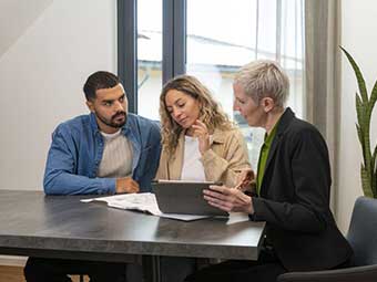 Foto: Drei Personen sitzen an einem Tisch, auf dem ein Gebäudeplan liegt und schauen darauf. Eine Person hält ein Tablet. 
