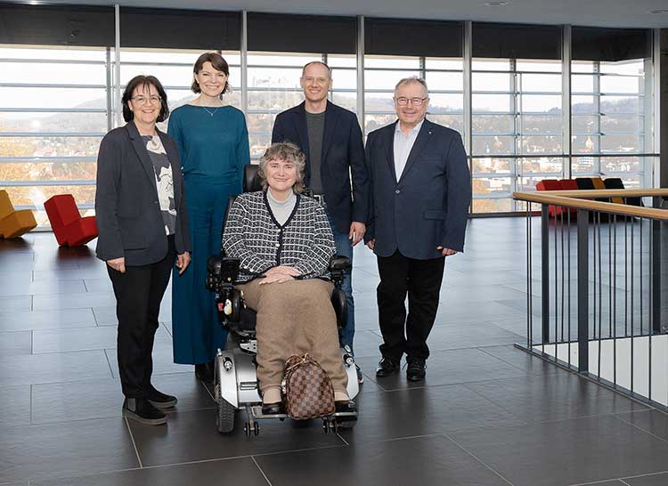 Foto: Fünf Personen in einem modernen Innenraum mit großen Fensterfronten. Eine Person sitzt vorne in einem elektrischen Rollstuhl. Die anderen vier Personen stehen dahinter und tragen überwiegend formelle Kleidung. 