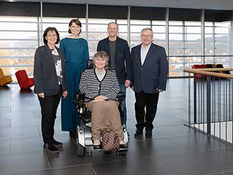 Foto: Fünf Personen in einem modernen Innenraum mit großen Fensterfronten. Eine Person sitzt vorne in einem elektrischen Rollstuhl. Die anderen vier Personen stehen dahinter und tragen überwiegend formelle Kleidung. 