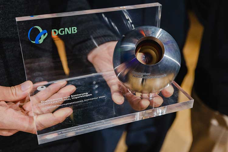 Trophäe aus Acrylglas mit dem Logo der DGNB und einer Gravur mit einer Auszeichnung für nachhaltige Architektur sowie einer Metallkugel mit einem Loch in der Mitte. Sie wird von zwei Händen präsentiert. 