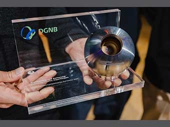 Trophäe aus Acrylglas mit dem Logo der DGNB und einer Gravur mit einer Auszeichnung für nachhaltige Architektur sowie einer Metallkugel mit einem Loch in der Mitte. Sie wird von zwei Händen präsentiert. 