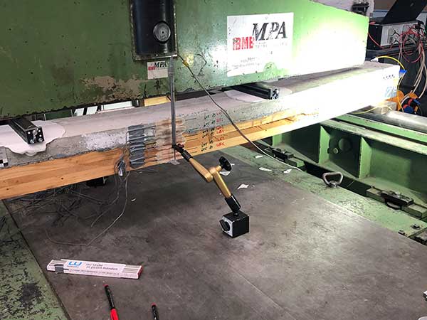 Foto: Vierpunkt-Biegeversuch an einer mittelgroßen Holz-Beton-Verbundplatte, an der mehrere Sensoren befestigt sind,  Die Platte liegt auf einer Testmaschine. 