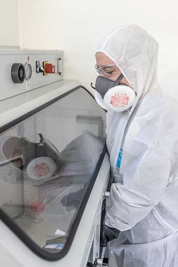 Foto: Person in Schutzkleidung und Atemschutzmaske in einem Gefahrstofflabor. Im Inneren einer Apparatur, hinter Glas, wird eine Materialprobe untersucht. 