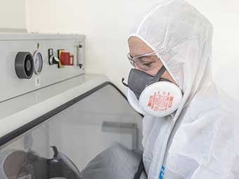 Foto: Person in Schutzkleidung und Atemschutzmaske in einem Gefahrstofflabor. Im Inneren einer Apparatur, hinter Glas, wird eine Materialprobe untersucht. 