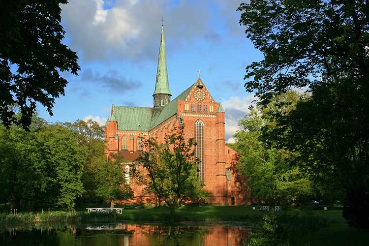 Foto: Gotische Backsteinkirche mit grünem Spitzdach und Turm im Zentrum, hohen, spitzbogigen Fenstern und Verzierungen, umgeben von Bäumen und Grünflächen. Im Vordergrund ein Teich. 
